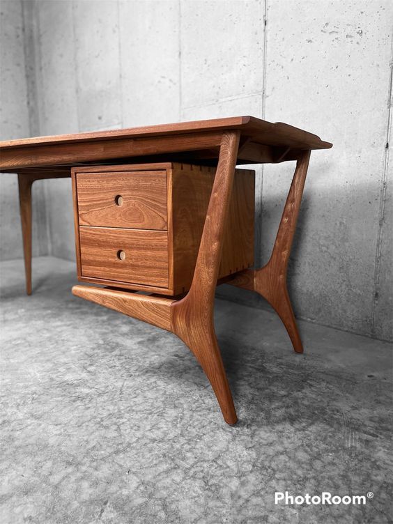 a wooden desk with two drawers on the top and one drawer at the bottom, in front of a concrete wall