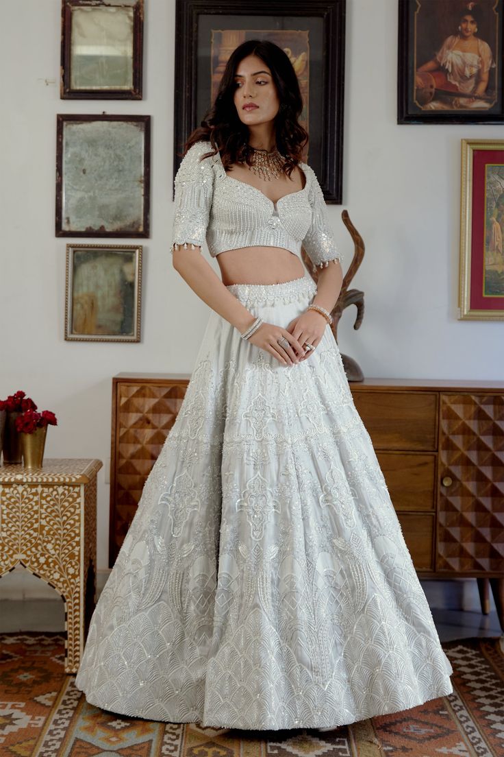 Divine is what comes to one’s mind while seeing up this moonlit beauty. Adorned with the purity of white tone and the hand-printed motifs embellished with white crystal beads and pearl moti sets up the ethereal mood for the celebrations. A perfect blend of modernity and tradition curated for the modern Indian brides.• Elbow-length blouse with a sweetheart neckline adorned with white pearl moti, crystal beads, and mirror-work. • Blouse contains a zip along with a metal hook on the left side.• The flowy translucent white embroidered dupatta with embroidered border. • The hand-printed motifs adorn the lehenga skirt embellished with handcrafted embroidery highlighting the motifs. • Lehenga contains a left zipper for support.From Moledro’s Fitoor collection. DELIVERY TIMEPlease allow 8-12 weeks Silver Lehenga With Pearl Embroidery For Reception, Traditional White Hand-embellished Lehenga, Elegant White Embellished Lehenga, Silver Sets With Pearl Embroidery For Party, Festive White Lehenga With Pearl Embroidery, Elegant White Lehenga With Mirror Work, Hand Embellished White Wedding Sets, Elegant White Festive Sets, Designer White Lehenga With Pearl Embroidery