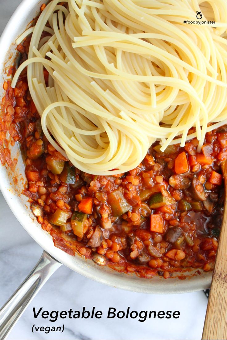 spaghetti and vegetables are being cooked in a skillet with a wooden spoon on the side