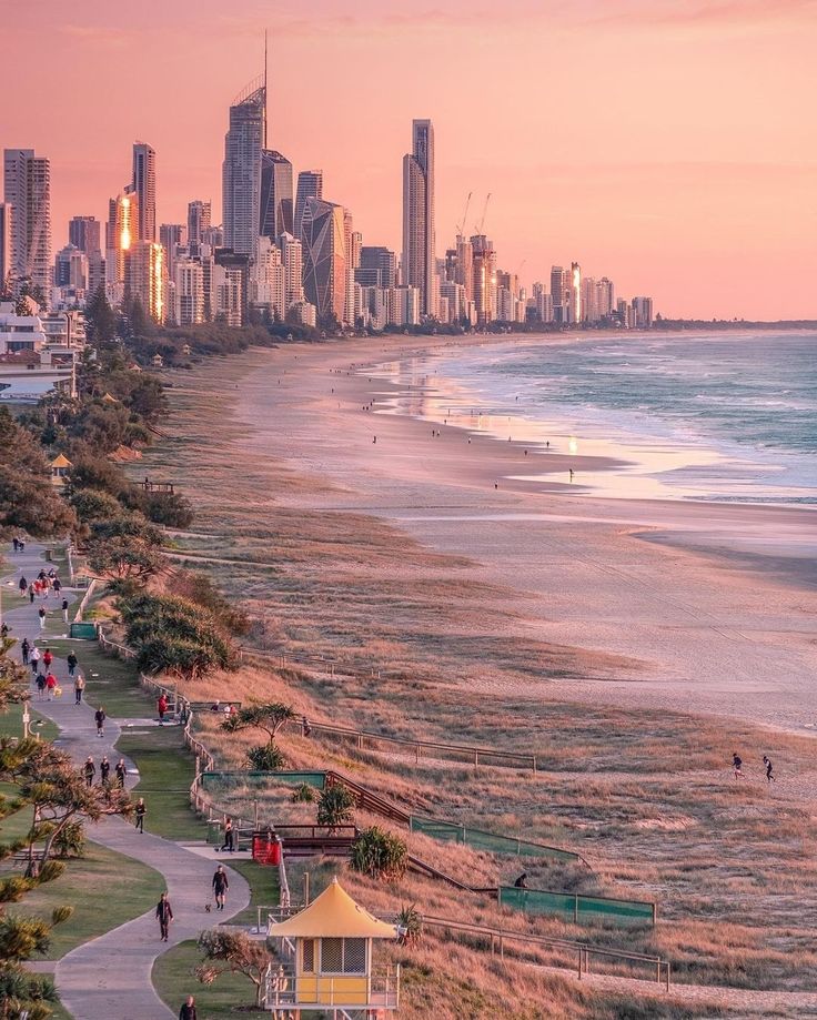 people are walking along the beach in front of some tall buildings and skyscrapers at sunset