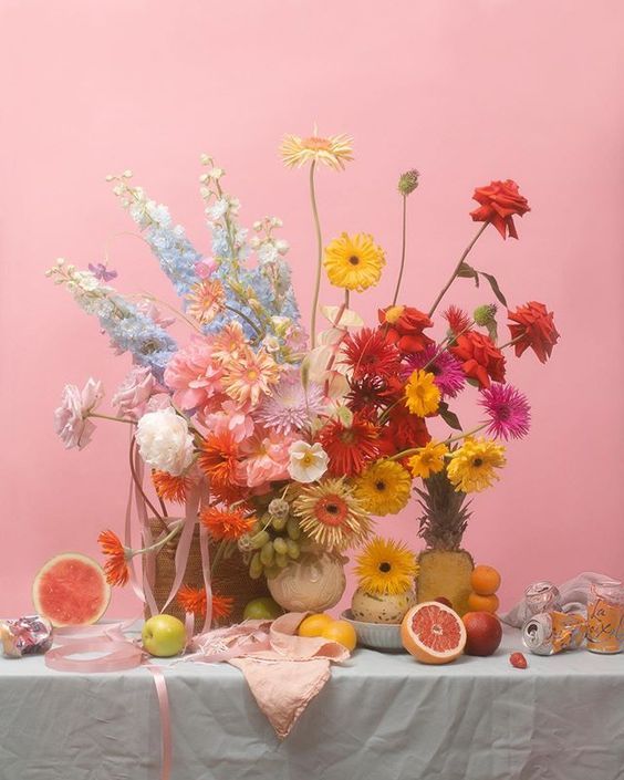 a table topped with lots of different types of flowers and fruit next to a pink wall