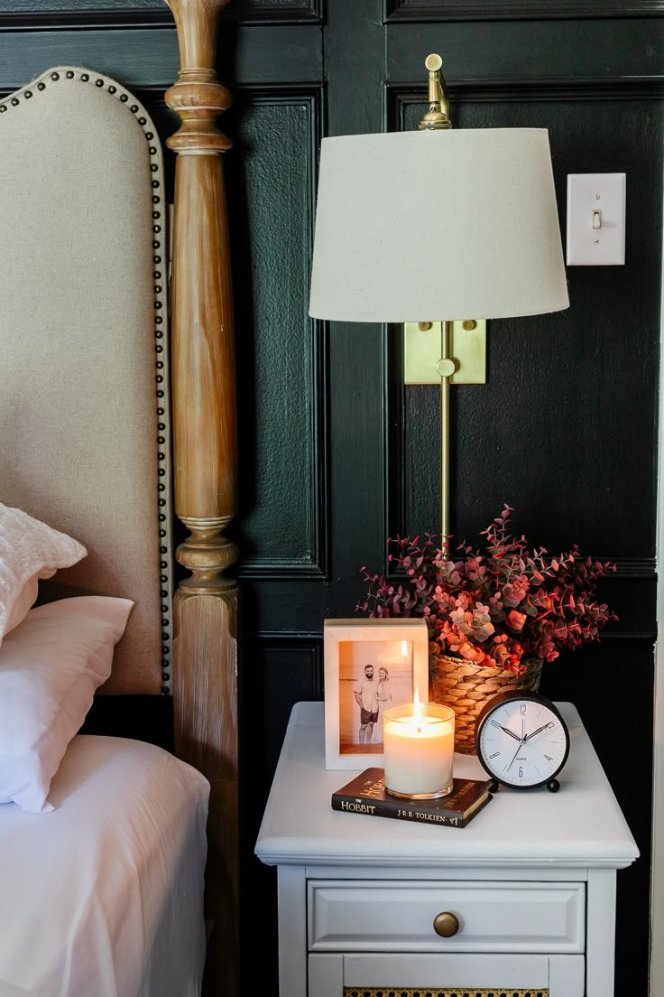 a nightstand with a clock, lamp and flowers on it next to a night stand