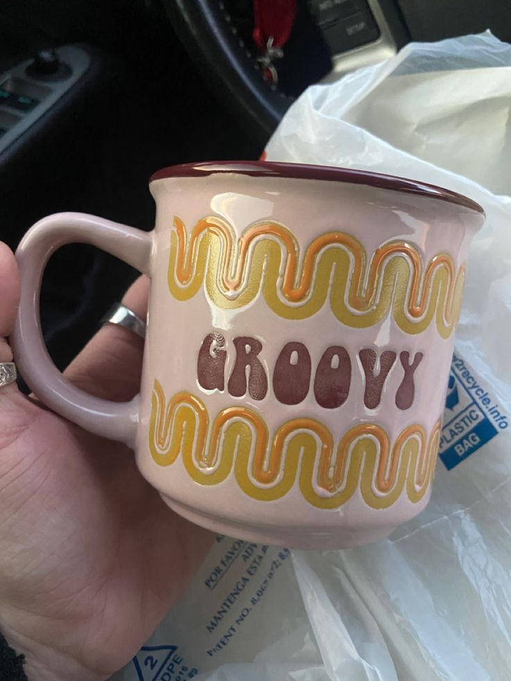 a hand holding a pink coffee mug with the words grooy on it in front of a car steering wheel