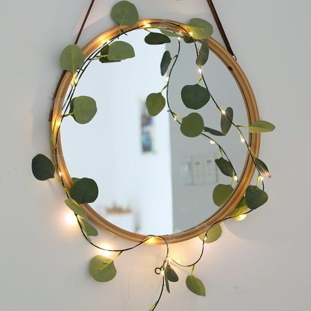 a round mirror hanging on the wall with some lights attached to it and green leaves