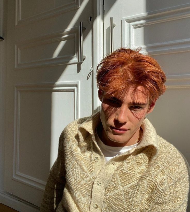 a young man with red hair wearing a sweater and looking at the camera while standing in front of a white door