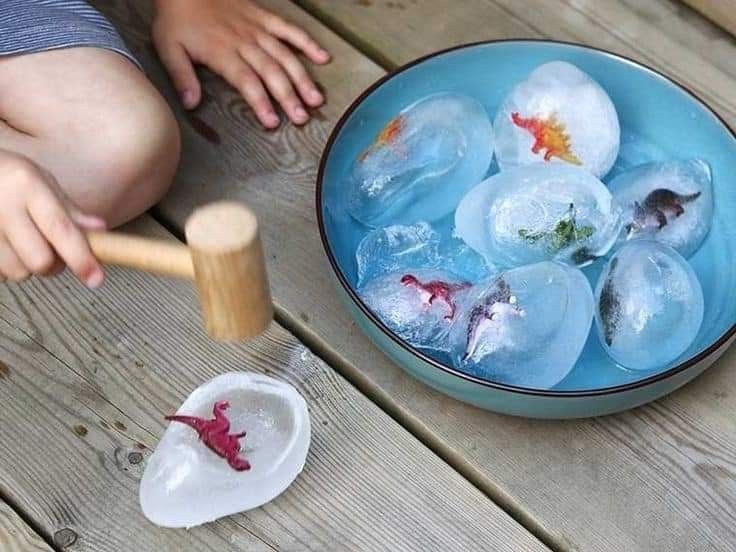 a child playing with ice cubes in a bowl on a wooden table next to a spoon