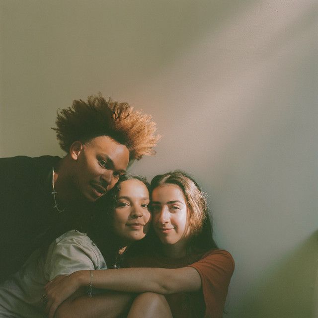 three young women hugging each other in front of a wall with sunlight coming through the window