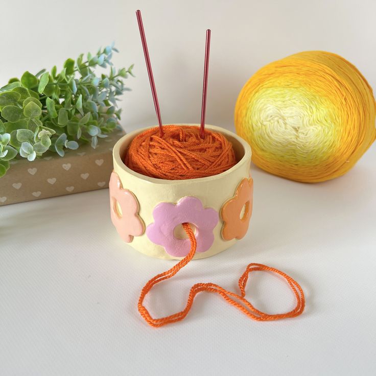 yarn and knitting needles sitting in a bowl next to an orange ball of yarn on a table