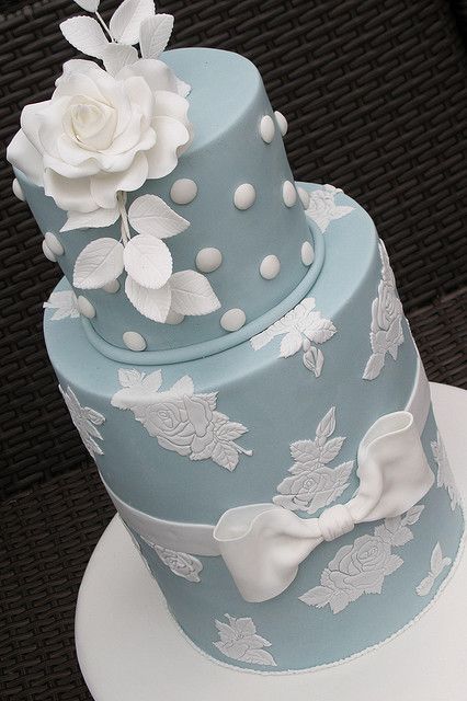 a three tiered blue cake with white flowers and bows on the top, sitting on a table