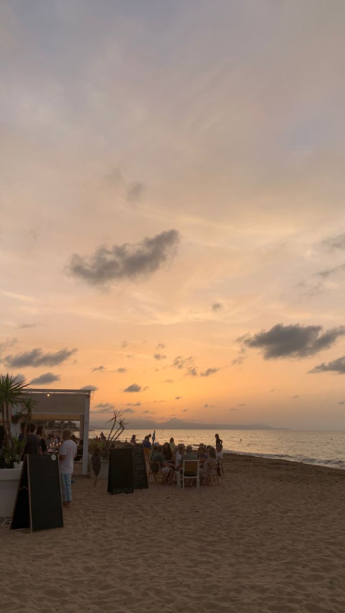 people sitting at tables on the beach watching the sun go down in the distance,