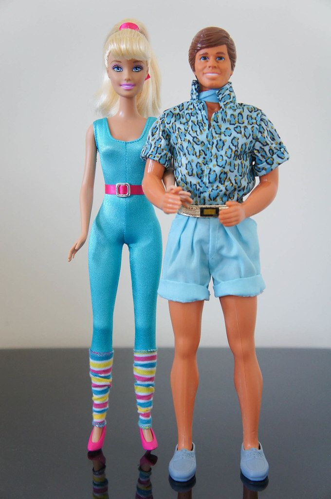 two barbie dolls standing next to each other on a black surface with one wearing blue and the other pink