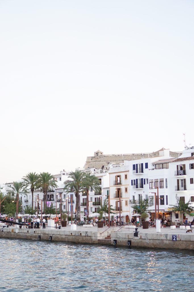 people are walking along the water in front of white buildings and palm trees on either side