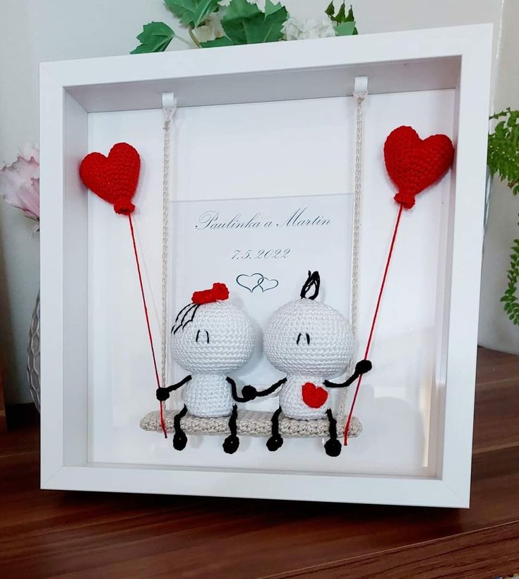 two crocheted hearts hanging from strings in a white frame on a wooden table