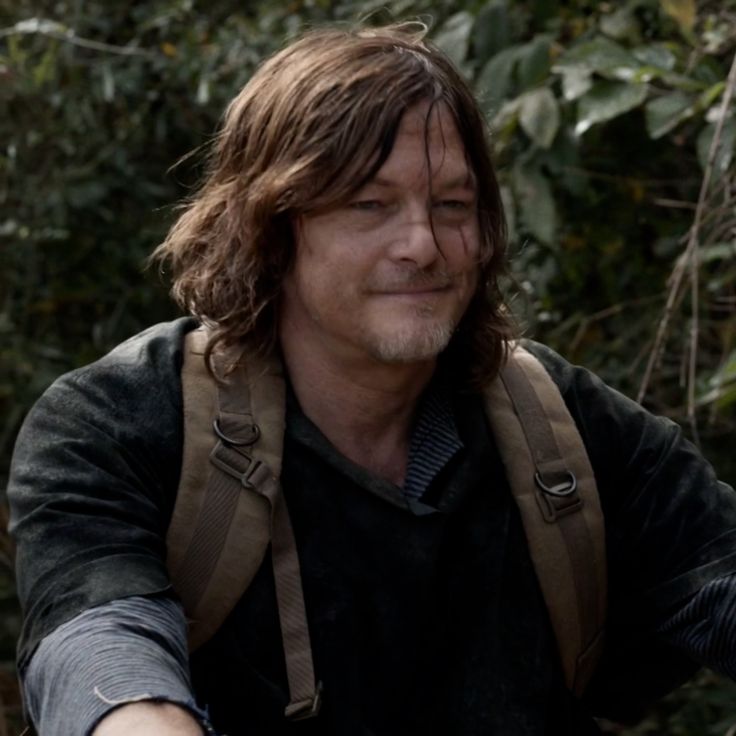 a man with long hair and backpacks in the woods pointing his hand at something