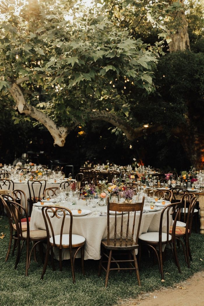 an outdoor dinner table set up with white linens and wooden chairs, surrounded by greenery