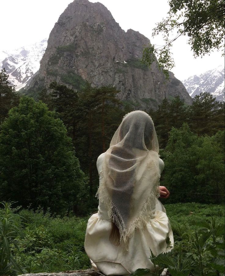 a woman in a white veil walking through the woods with mountains in the back ground