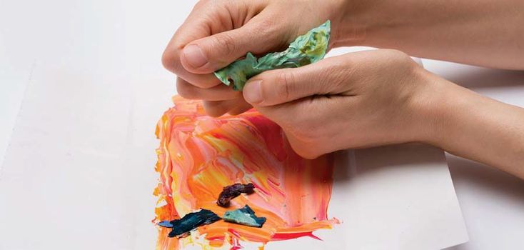 two hands are painting on paper with orange and green colors while another hand is holding something in their other hand