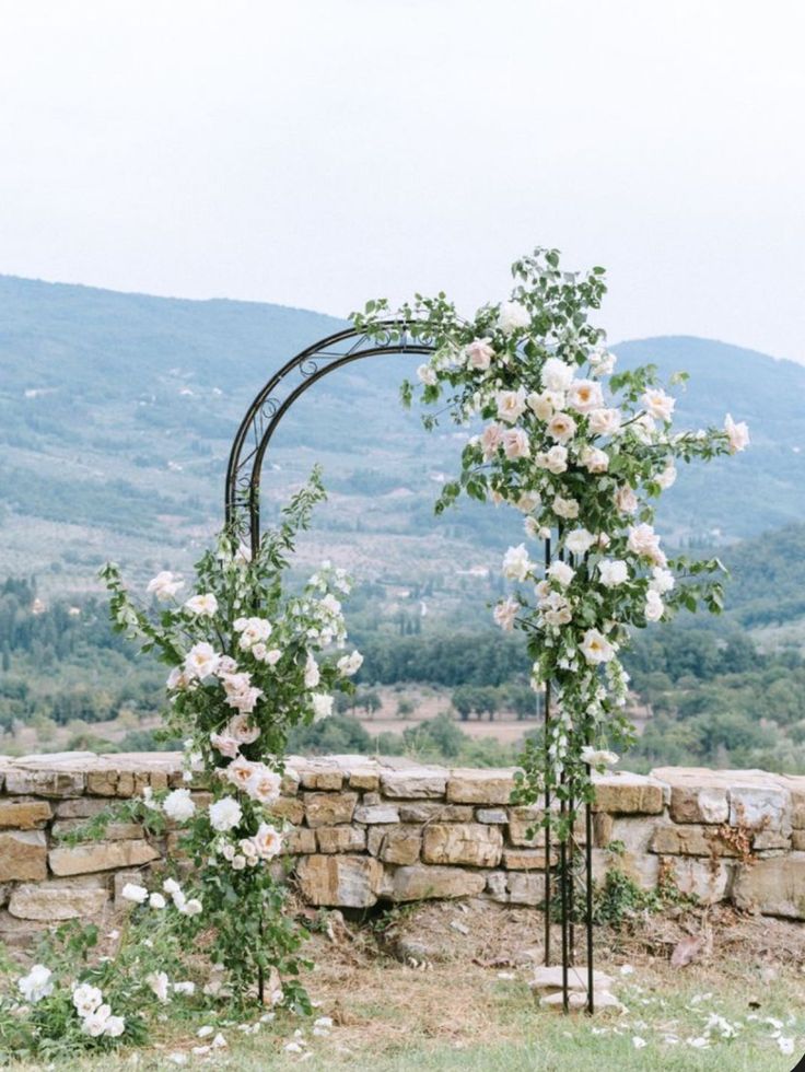 a wedding arch with white flowers on it in front of a stone wall and mountains