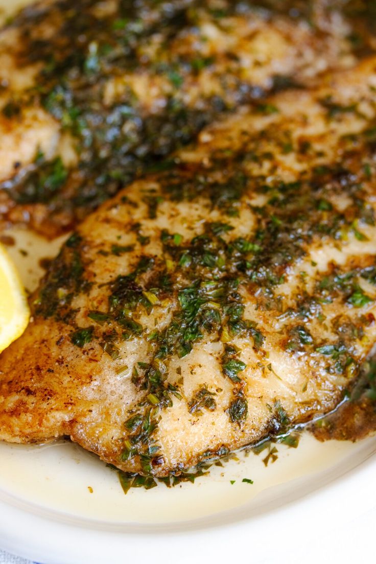 a white plate topped with meat covered in herbs and lemon wedges next to a slice of lemon