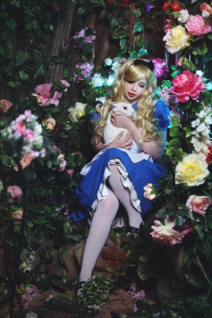 a woman in a blue dress is holding a white cat surrounded by flowers and greenery