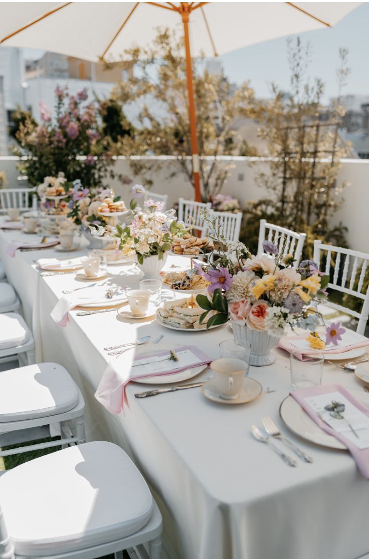 an outdoor table set up with white chairs and pink napkins, plates and utensils