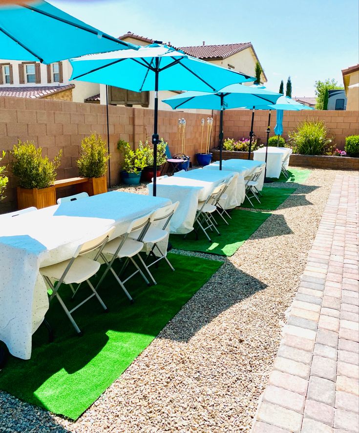 a long table with blue umbrellas is set up in the yard for an event