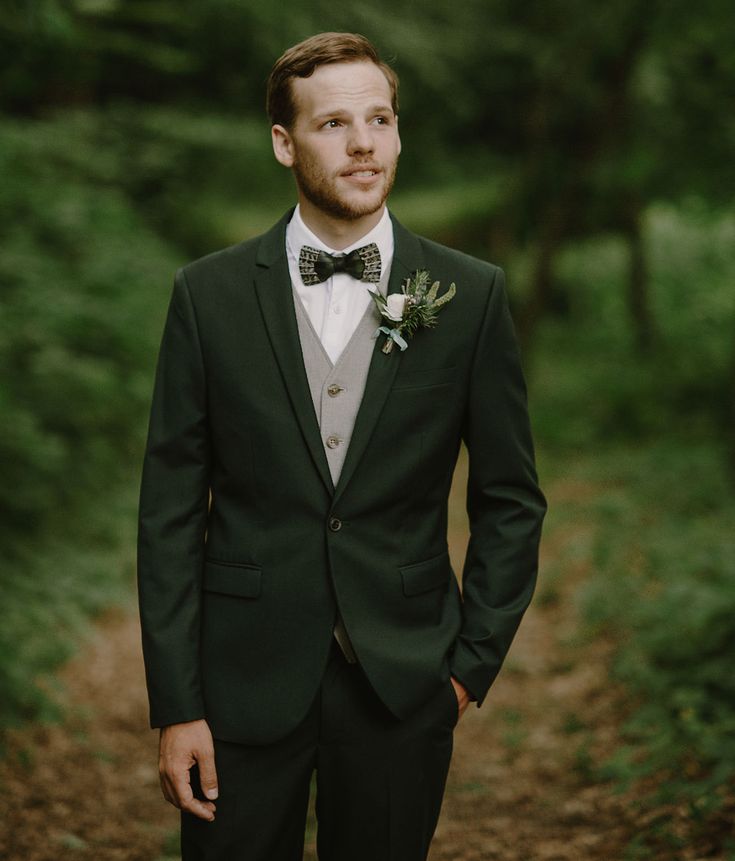 a man in a suit and bow tie standing on a dirt road with trees behind him