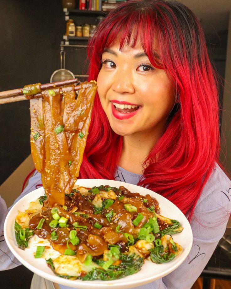 a woman with red hair holding a plate of food and chopsticks in her mouth