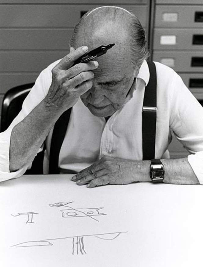 an older man sitting at a table with his hand on his head and writing on the paper