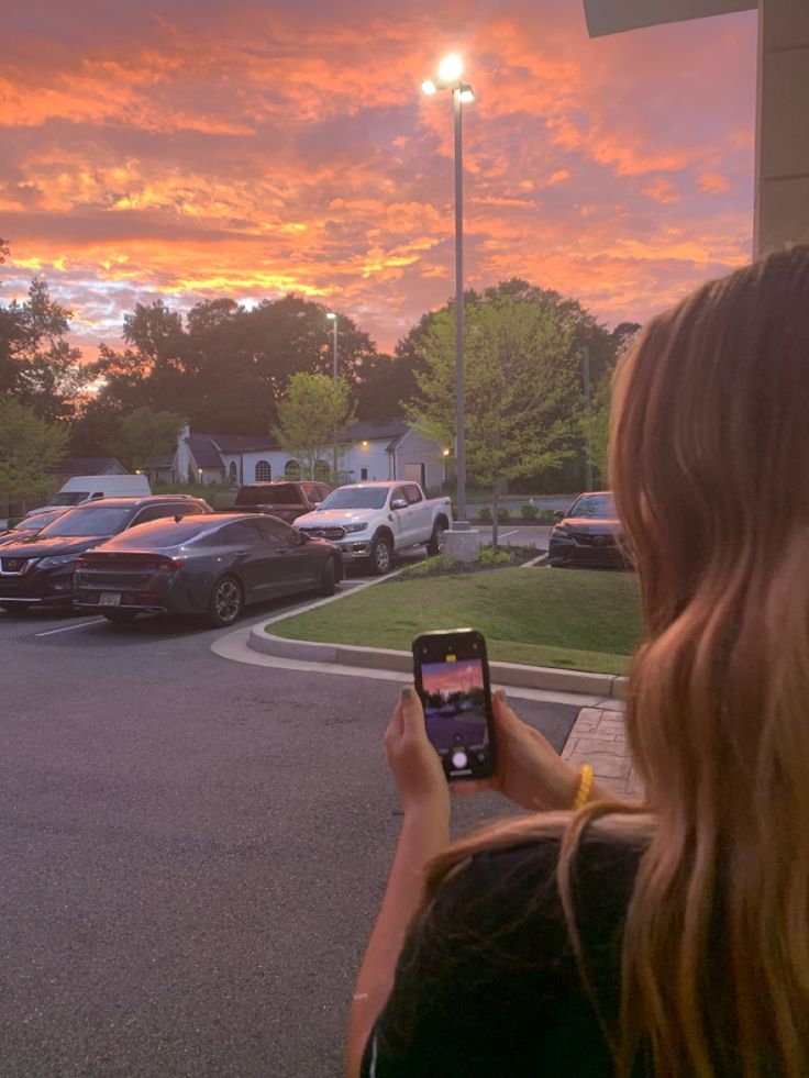 a woman holding up her cell phone to take a picture of the sunset in front of some parked cars