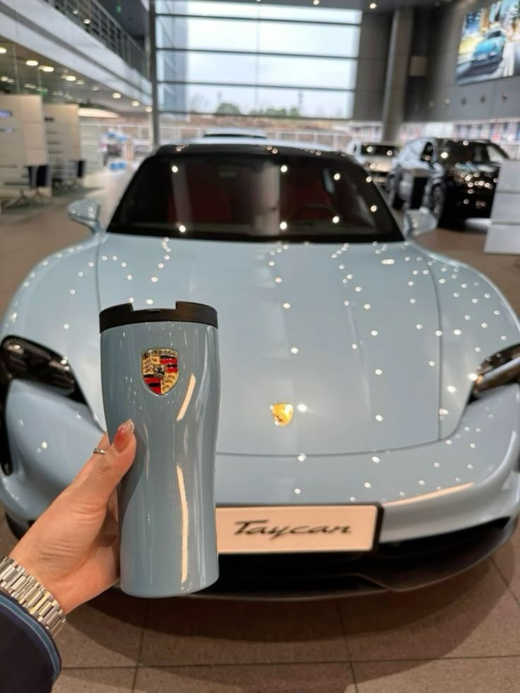 a person holding up a cup in front of a blue sports car at an auto show