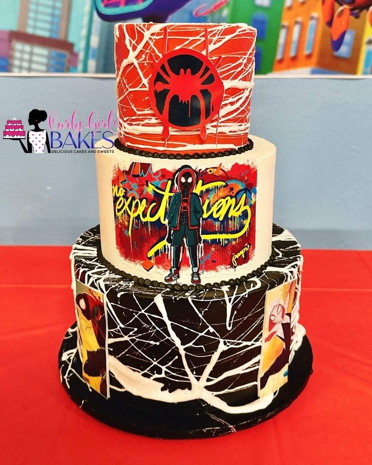 a three tiered cake with spiderman decorations on the top and bottom, sitting on a red table cloth