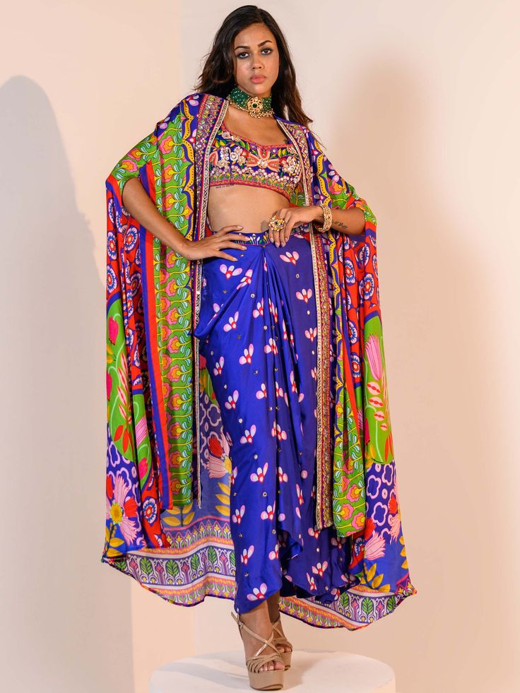 A three-piece Blue Indica Mirror Dhoti Skirt Set Paired with Bralette and Cape from the Juhi Bengani collection – Rang-Festive 2022. This gorgeous blue indica silk mirror dhoti skirt is paired with a heavy embroidered multicolored bralette blouse. The bright and vibrant colors with floral print, heavy embroidery of mirror, beads, stones, and thread sequence work enhance this satin dhoti skirt set. The indica blue dhoti skirt has a side opening at the waistline. This outfit is completed with a Bollywood Style Silk Palazzo Set For Navratri, Navratri Silk Palazzo Set With Traditional Drape, Designer Blue Silk Sharara, Blue Bollywood Art Silk Palazzo Set, Blue Silk Sets For Diwali, Blue Silk Set For Diwali, Blue Silk Set For Navratri, Festive Blue Art Silk Palazzo Set, Blue Palazzo Set For Reception And Festivals