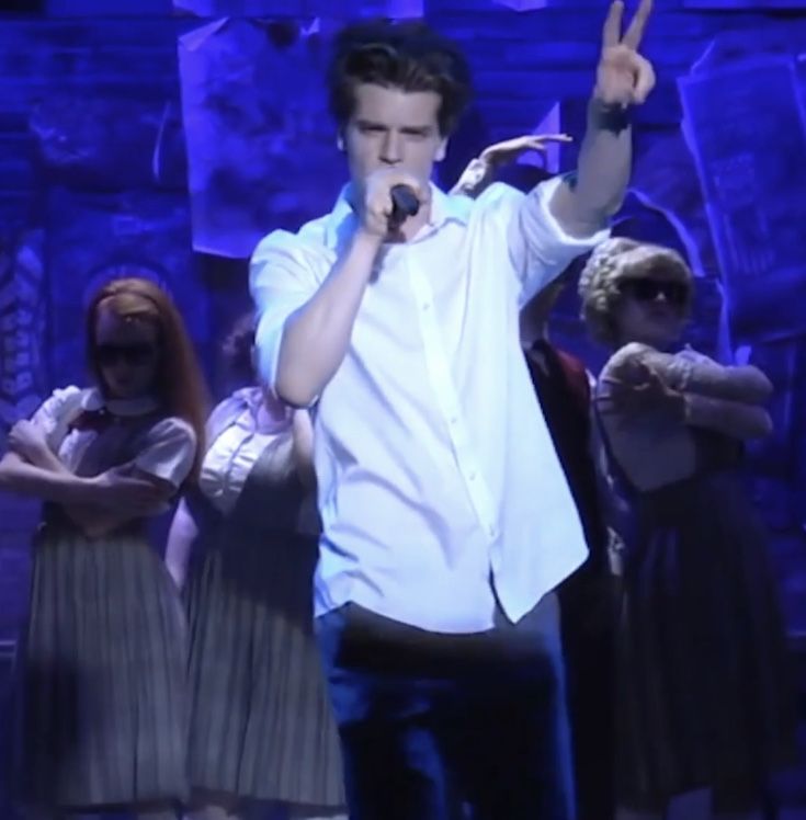 a man in white shirt and blue pants on stage with other people behind him holding up their hands