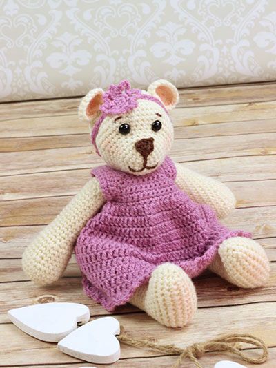 a crocheted teddy bear in a purple dress sitting on a wooden floor next to a string