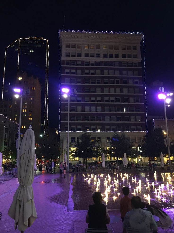 people are sitting in the middle of a fountain with lights on it and buildings in the background