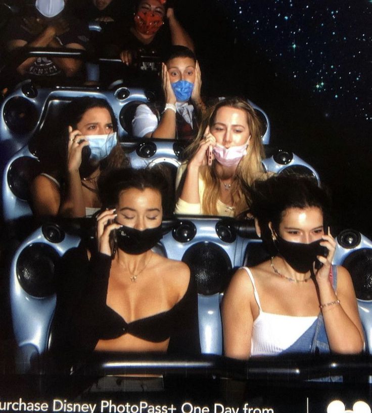 several women wearing face masks ride in a roller coaster at an amusement park, while one woman covers her mouth