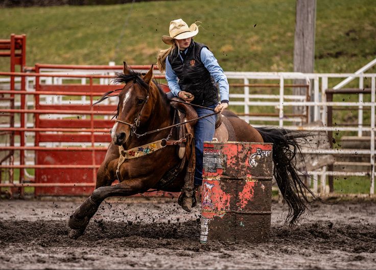 a woman riding on the back of a brown horse next to a red barrel in an arena