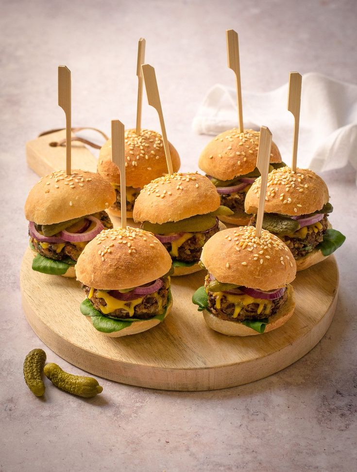 six hamburgers with toothpicks stuck in them on a cutting board next to pickles