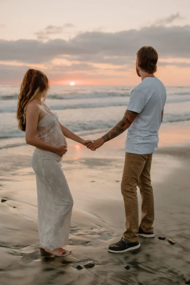 a man and woman holding hands while standing on top of a beach next to the ocean