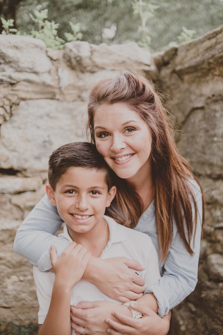 a woman holding a boy in her arms and smiling at the camera while standing next to a rock wall