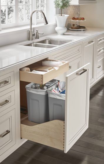 an open kitchen cabinet with two trash cans in the bottom drawer and a sink below