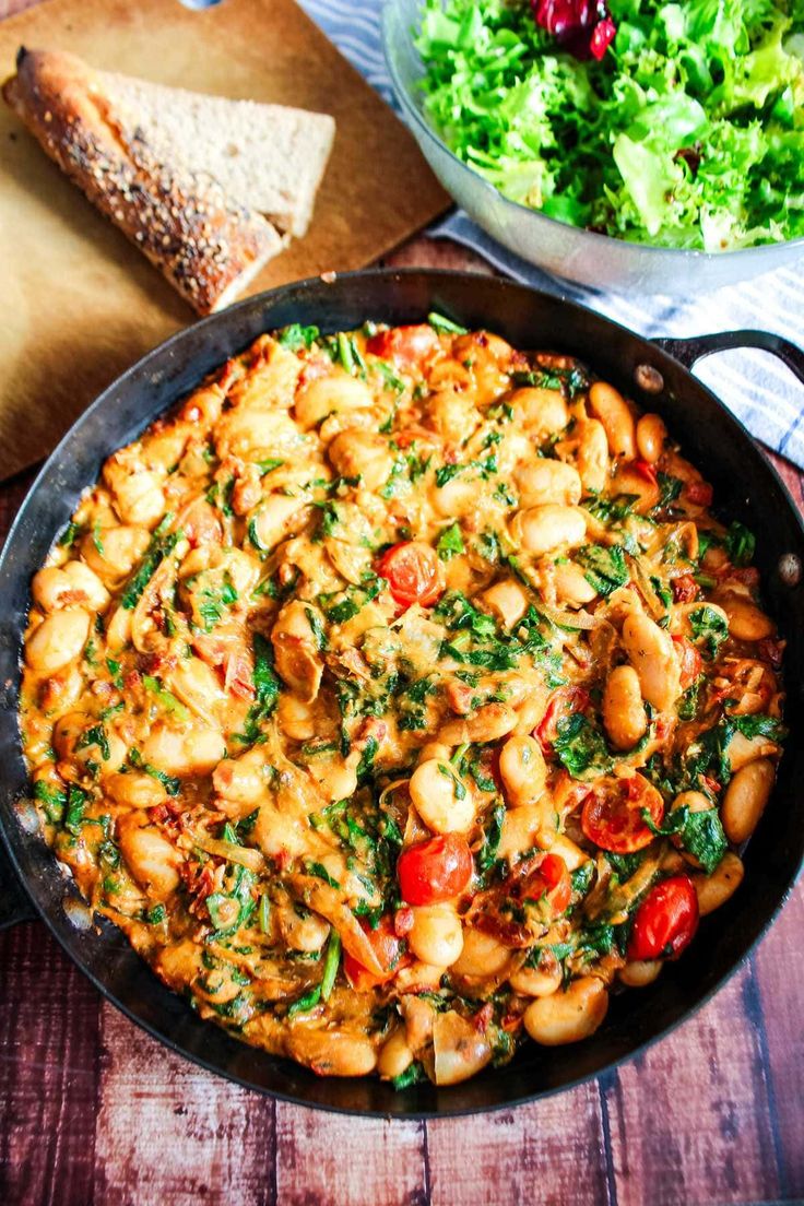 a skillet filled with pasta and vegetables next to a bowl of bread on a wooden table