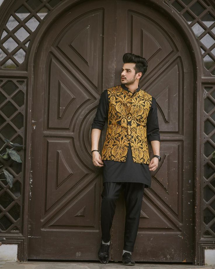 a man standing in front of a wooden door wearing a black and gold outfit with an intricate design on it