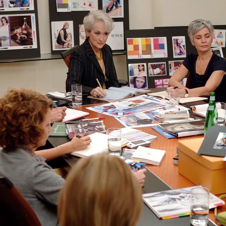 three women sitting at a table with books and magazines on it, surrounded by other people