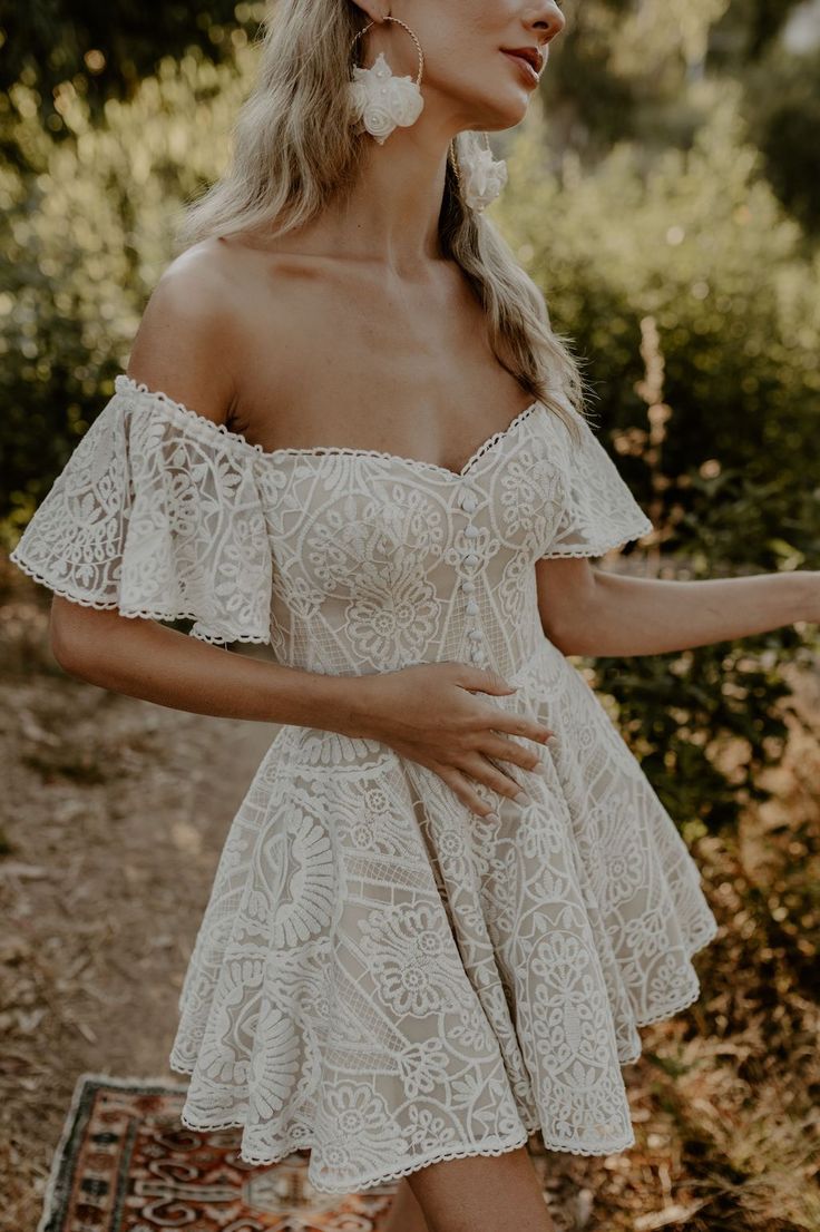 a woman in a white dress is standing near some bushes and trees with her hand on her hip