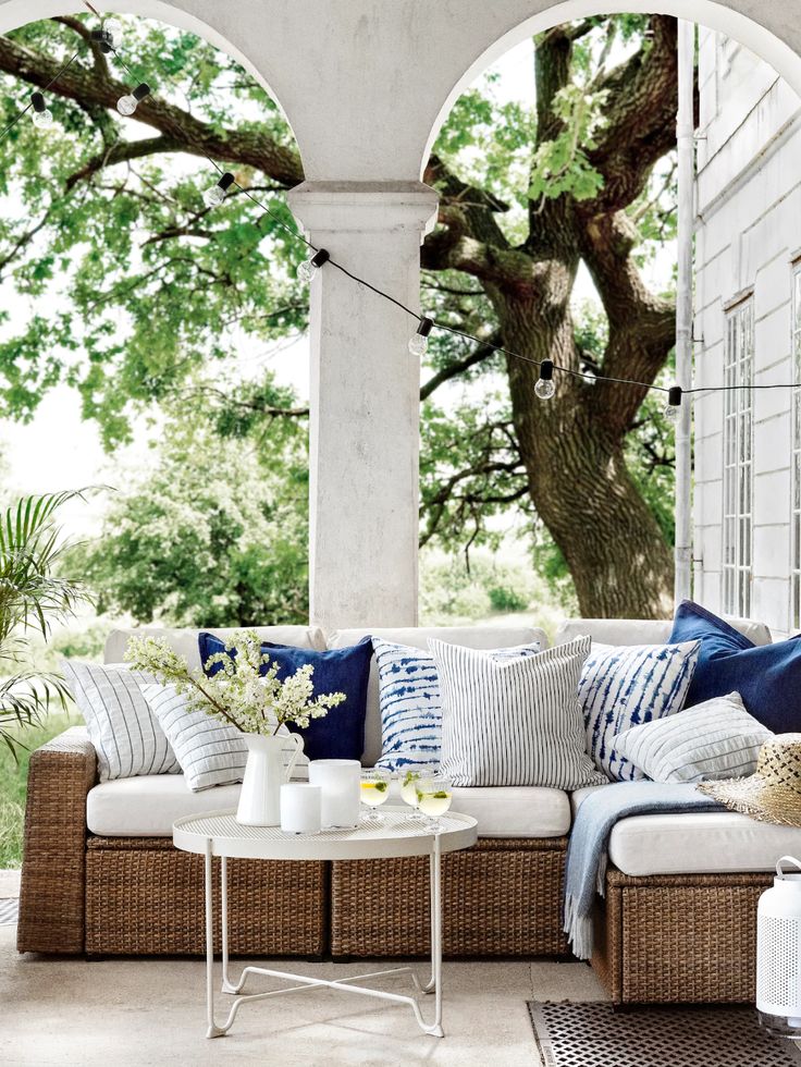 an outdoor living room with wicker furniture and blue pillows on the couch, along with large trees