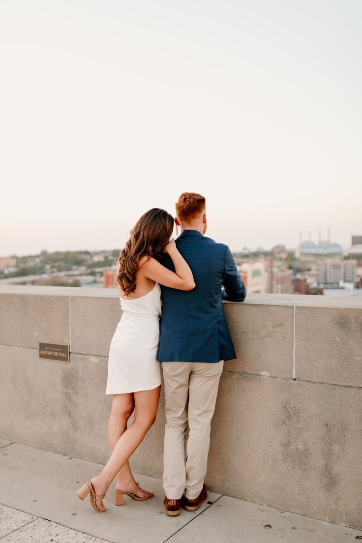 a man and woman leaning against a wall looking at each other with the city in the background