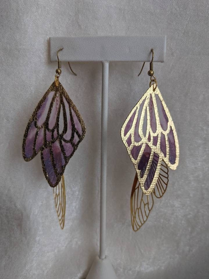 These unique and whimsical earrings feature gorgeous colored and golden faerie wings. Perfect for special occasions or to add a bit of magic to your everyday! Choose primary color between lavender/purple, aqua/sapphire, teal/purple, or purple/red! Gold Fantasy Handmade Earrings, Handmade Gold Fantasy Earrings, Gold Handmade Fantasy Earrings, Handmade Gold Wing-shaped Earrings, Purple Butterfly Earrings For Gift, Whimsical Purple Nickel-free Earrings, Gold Fairycore Drop Earrings, Fairycore Gold Drop Earrings, Gold Fairy Earrings For Gift