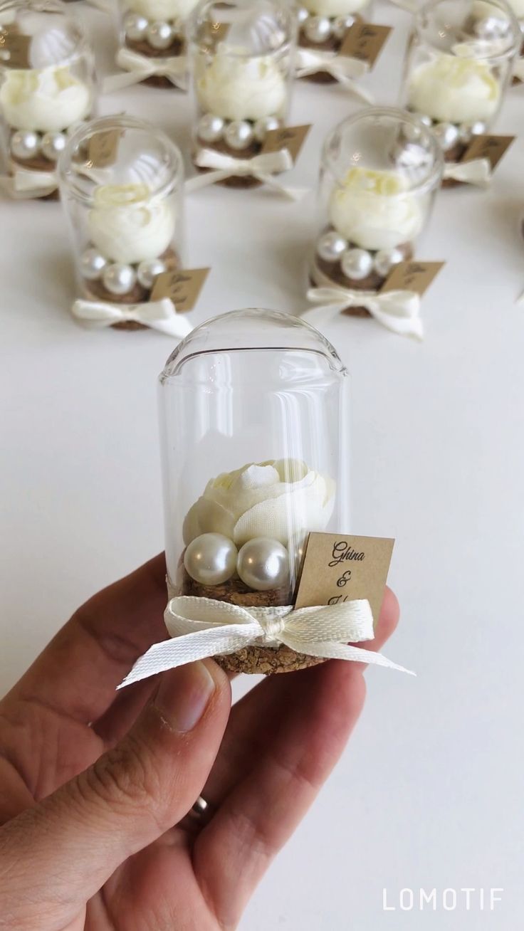 a hand holding a glass jar filled with white flowers and pearls on top of a table